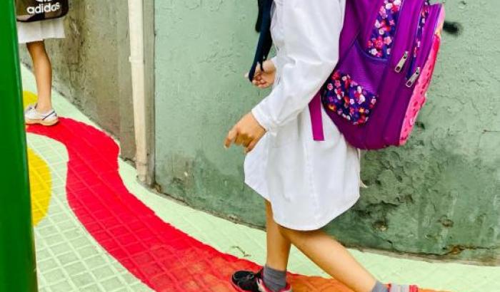 Niño escolar, de túnica, moña y mochila, camina por un zig zag pingado de rojo sobre un fondo verde en la vereda