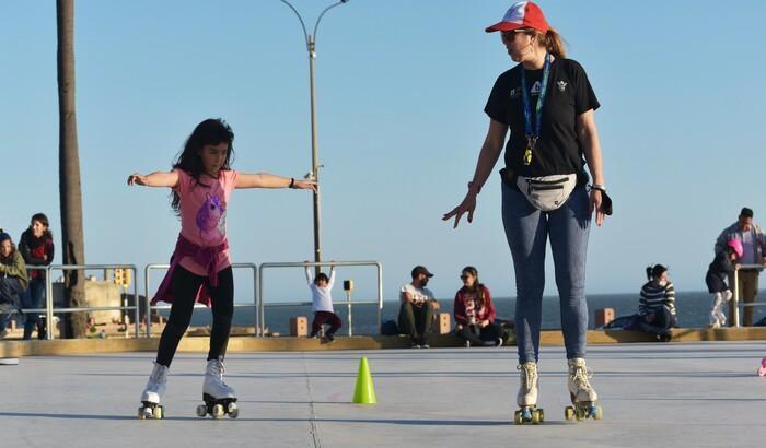 Fotografía en la que se puede ver: una niña patina junto a una profesora. La niña, de brazos abiertos, intenta rodear unos conos a modo de obstáculos